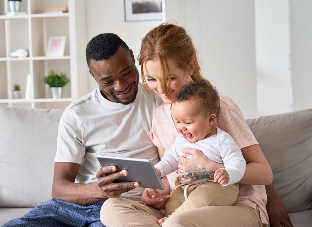 Personal Insurance - Happy Young Multiracial Family Couple Holding Cute Infant Daughter Having Fun With Digital Tablet Sitting on Sofa